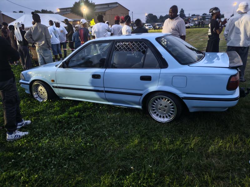 Side view of the light-blue Corolla on the grass with a crowd of people behind it