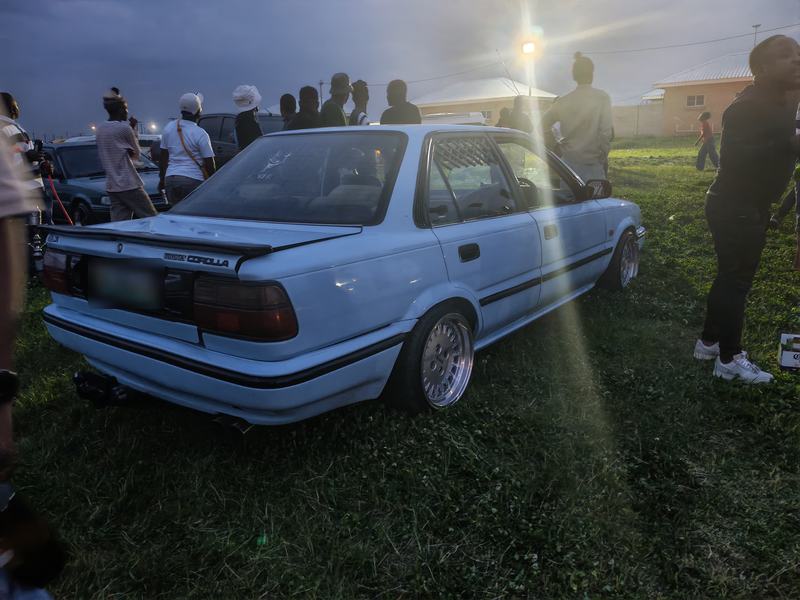 Rear three-quarter view of a lowered light-blue Toyota Corolla sedan on mesh wheels, sun flaring behind it