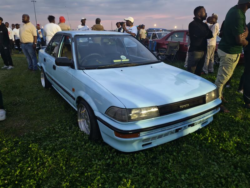 Front three-quarter view of the light-blue Corolla, people standing around it at dusk