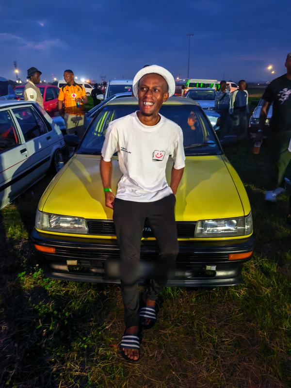 A man in a white bucket hat sits on the bonnet of a yellow Toyota at dusk, with the meet crowd behind him