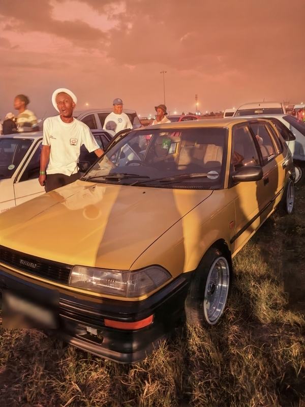 A gold Toyota hatchback on polished wheels under a pink sunset sky, with its owner behind it