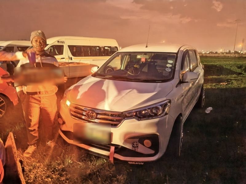 A man holds up a number plate next to his white Toyota Rumion at night