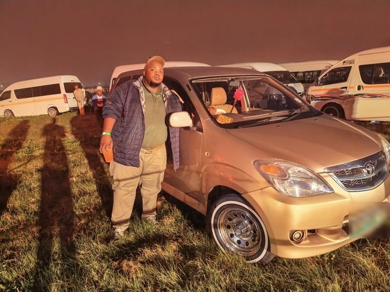 A man stands beside his gold Toyota Avanza on white-wall tyres, taxis parked behind
